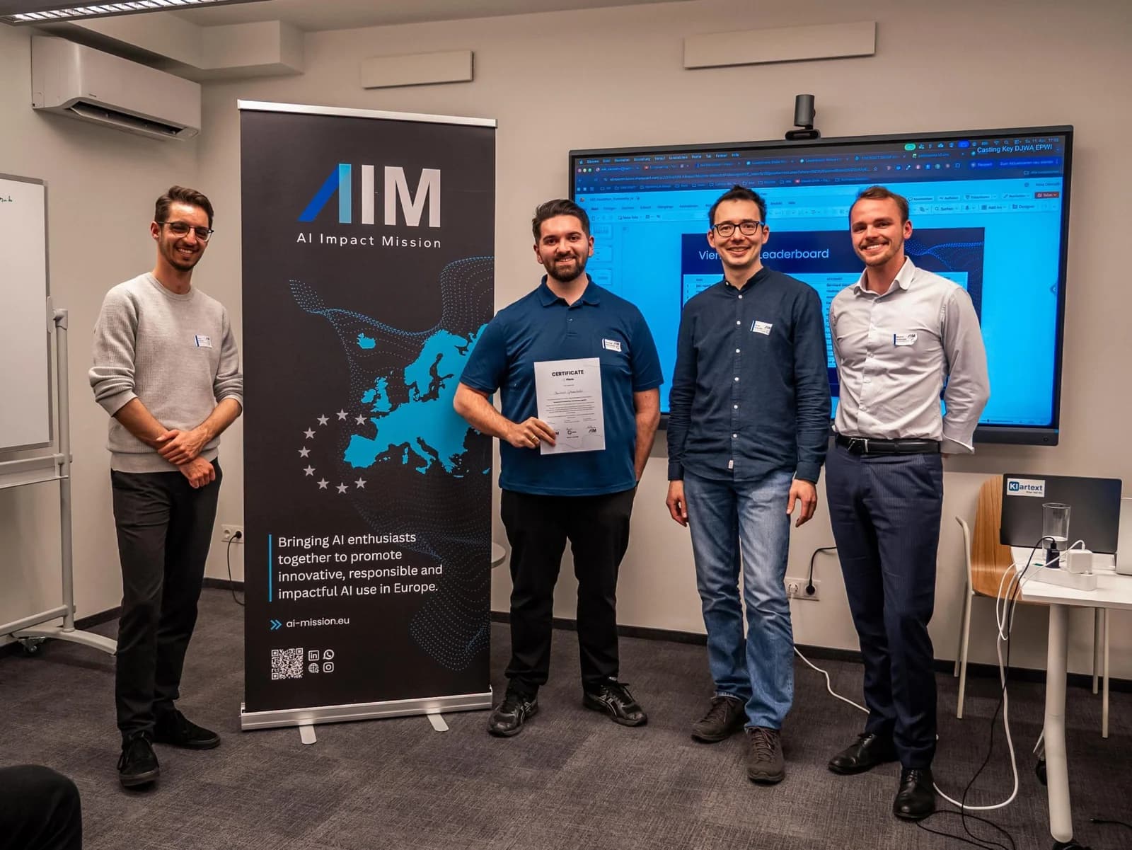 Vier Männer nebeneinander vor einem Rollup der AI Impact Mission. Bernhard Götzendorfer, Zweiter von links im blauen Polohemd, hält die Urkunde des BitGN PAC Hackathon.