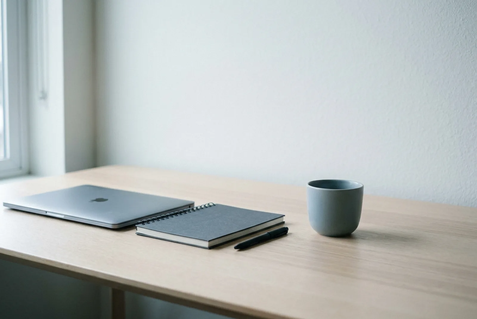 Minimalistischer Arbeitsplatz mit Laptop, Notizbuch und Tasse bei kühlem Tageslicht